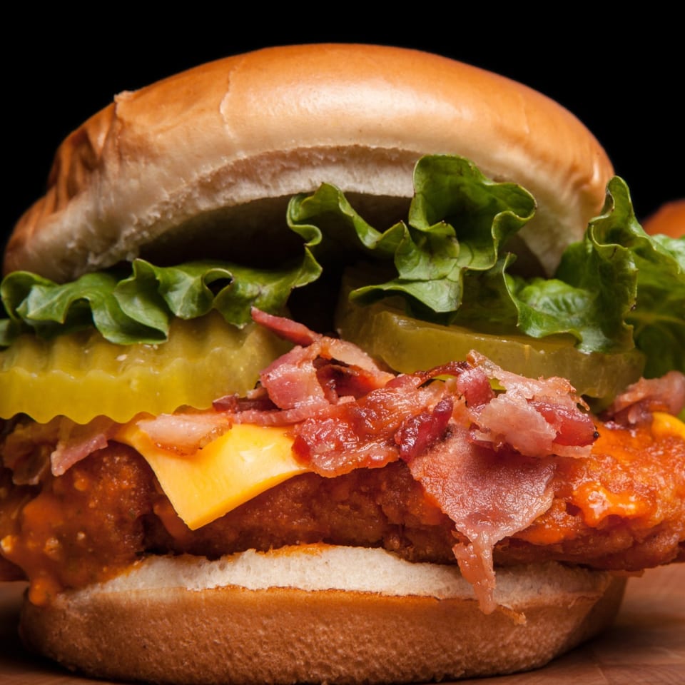 Close-up of a chicken sandwich with bacon, cheese, pickles, and lettuce on a bun.