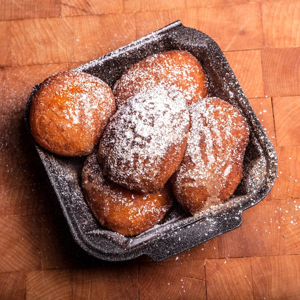 Five powdered sugar-dusted fried dough balls in a black container on a wooden surface.